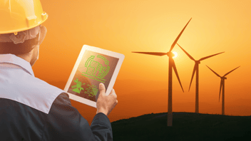 Engineer with hard hat, back to the viewer, holding a tablet against a backdrop of a wind farm at sunset